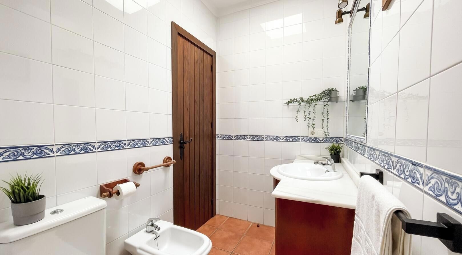 A bathroom with white tiled walls, a wooden door, a white sink, a bidet, and a mirror.