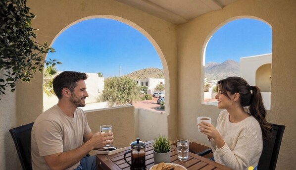 Two people sitting at a table with drinks, a coffee pot, and a small plant.