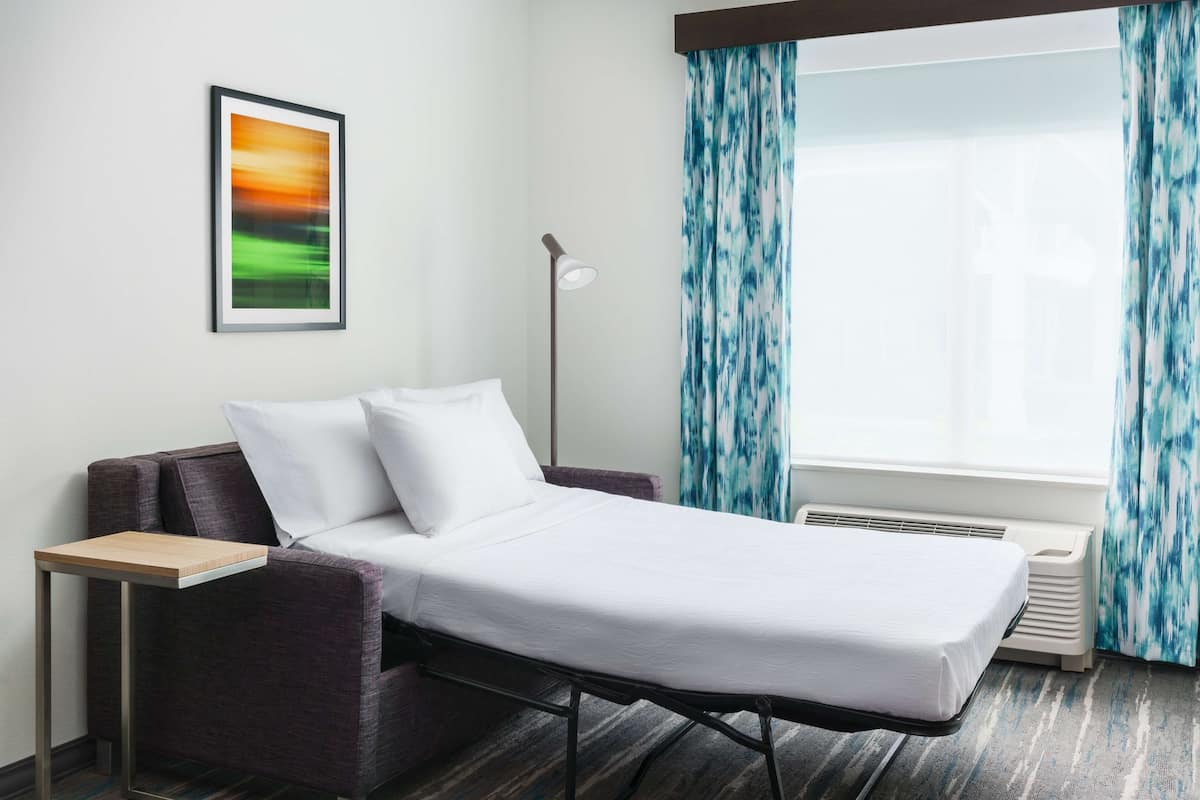 Een hotelkamer met een bed, een kleine tafel, een bank, een staande lamp en een raam met gordijnen.