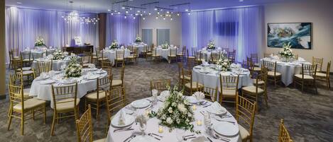 Een feestzaal met ronde tafels gedekt voor een formeel evenement, met witte tafelkleden, gouden stoelen en bloemstukken als decoratie.