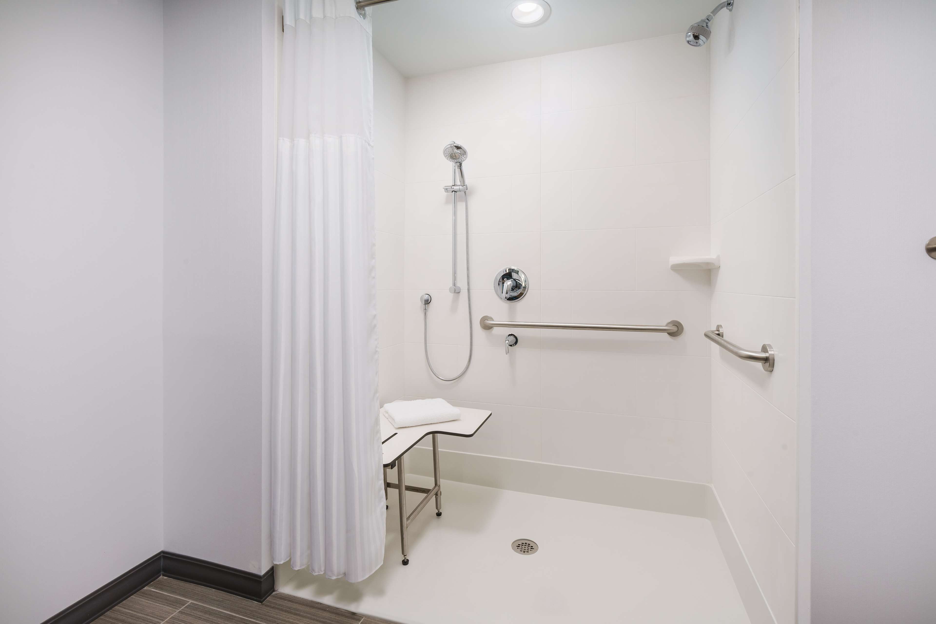 Un baño moderno con ducha, barras de apoyo, un pequeño banco y suelo de azulejos blancos.