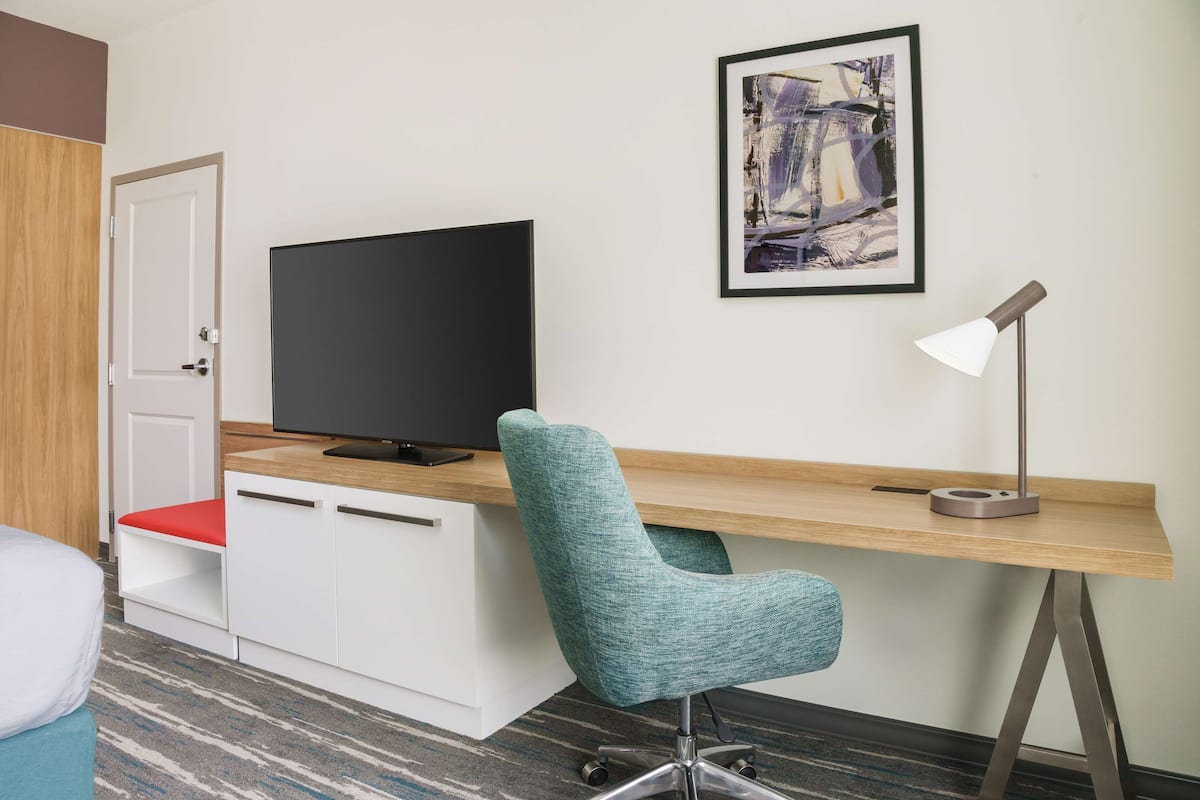 Een hotelkamer met een houten bureau, een blauwgroene bureaustoel, een flatscreen televisie, een witte nachtkastje en een rode kussen.