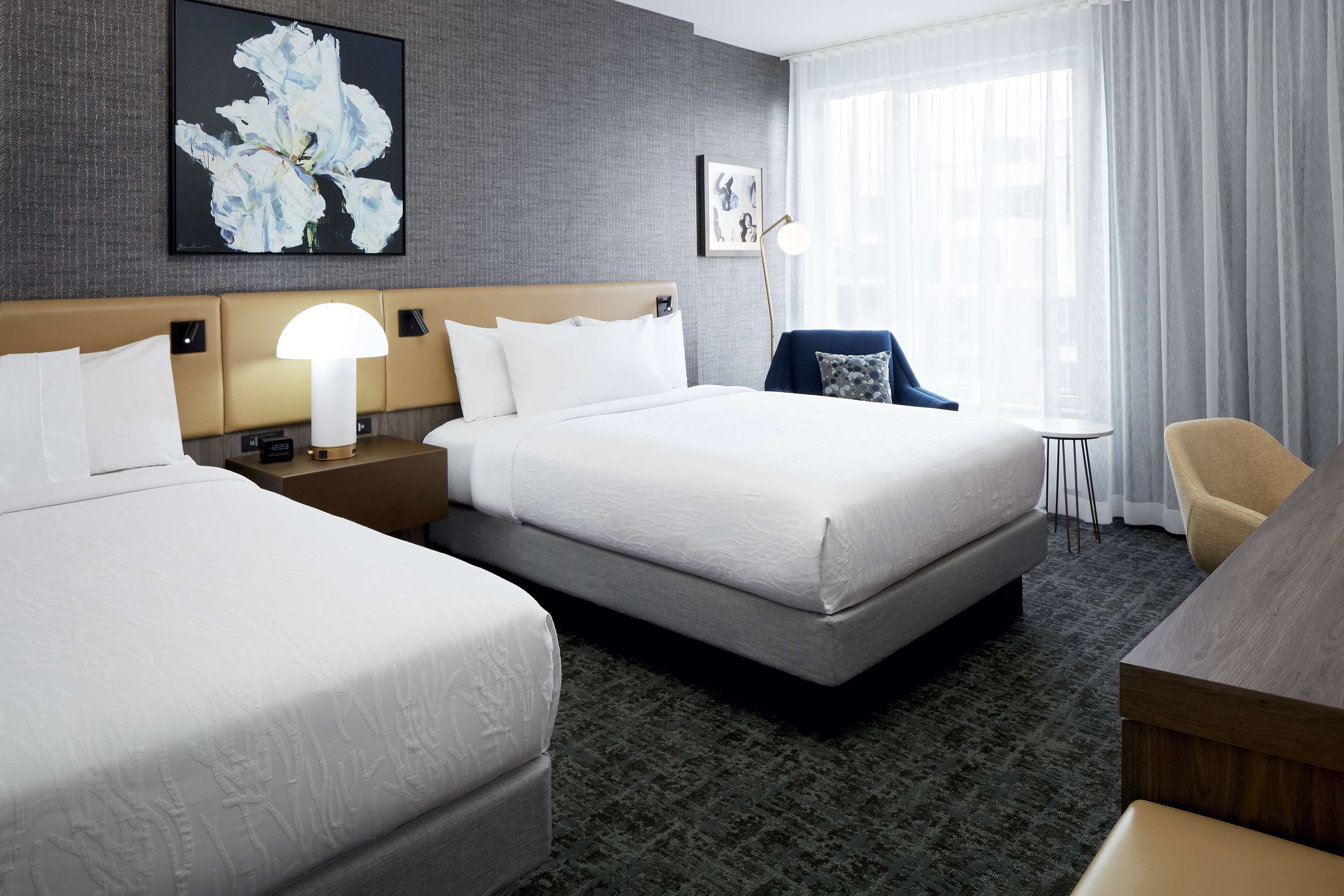 Quarto de hotel com duas camas, uma escrivaninha, uma cadeira e um abajur.