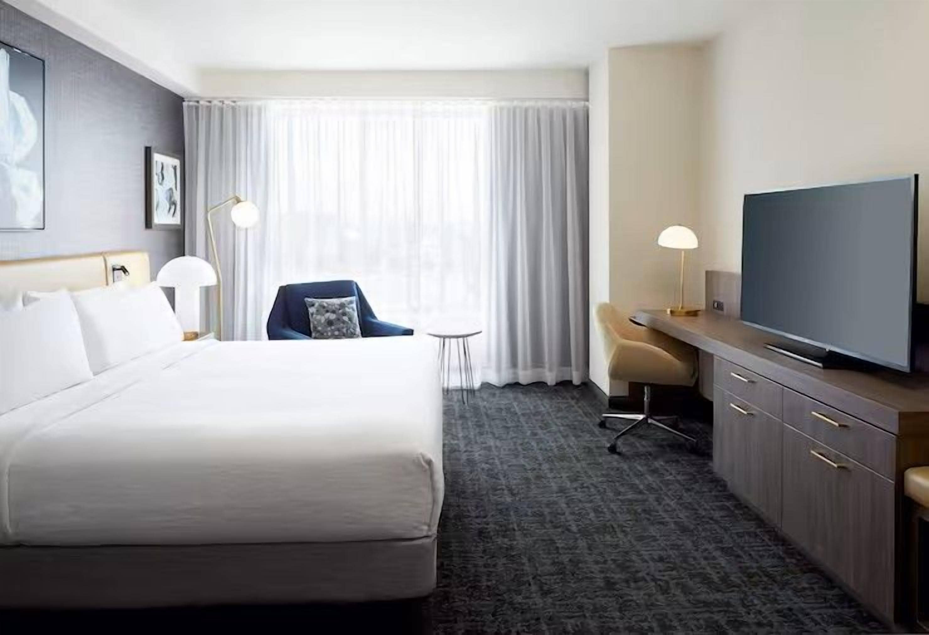 Ein Hotelzimmer mit einem großen Bett, einem Schreibtisch mit Flachbildfernseher, einem Sessel, einem kleinen Tisch und einer Lampe.