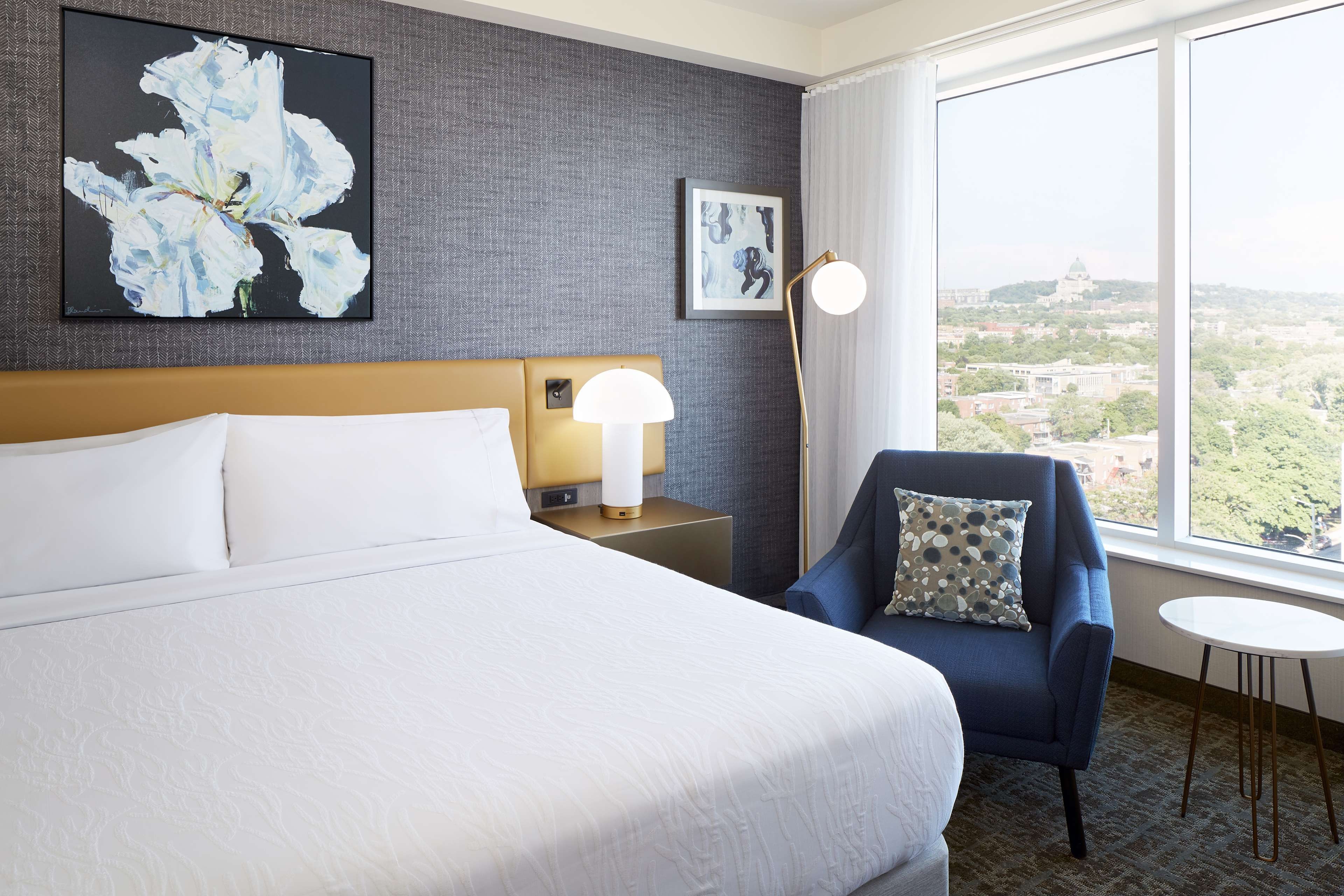 A hotel room with a large bed, a chair, a small table, a lamp, and a view of the city.