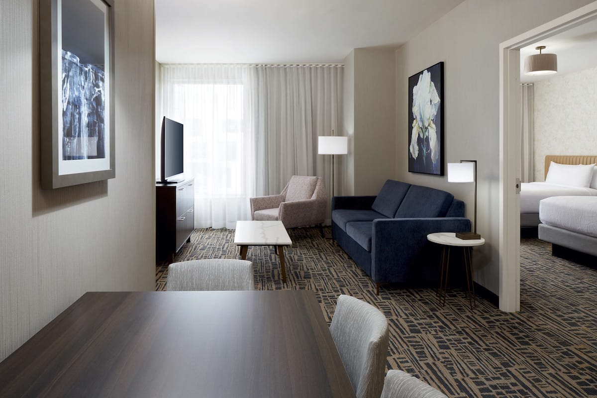 A hotel room with a dark wood table, a sofa, a chair, a TV, a lamp, and a bed.