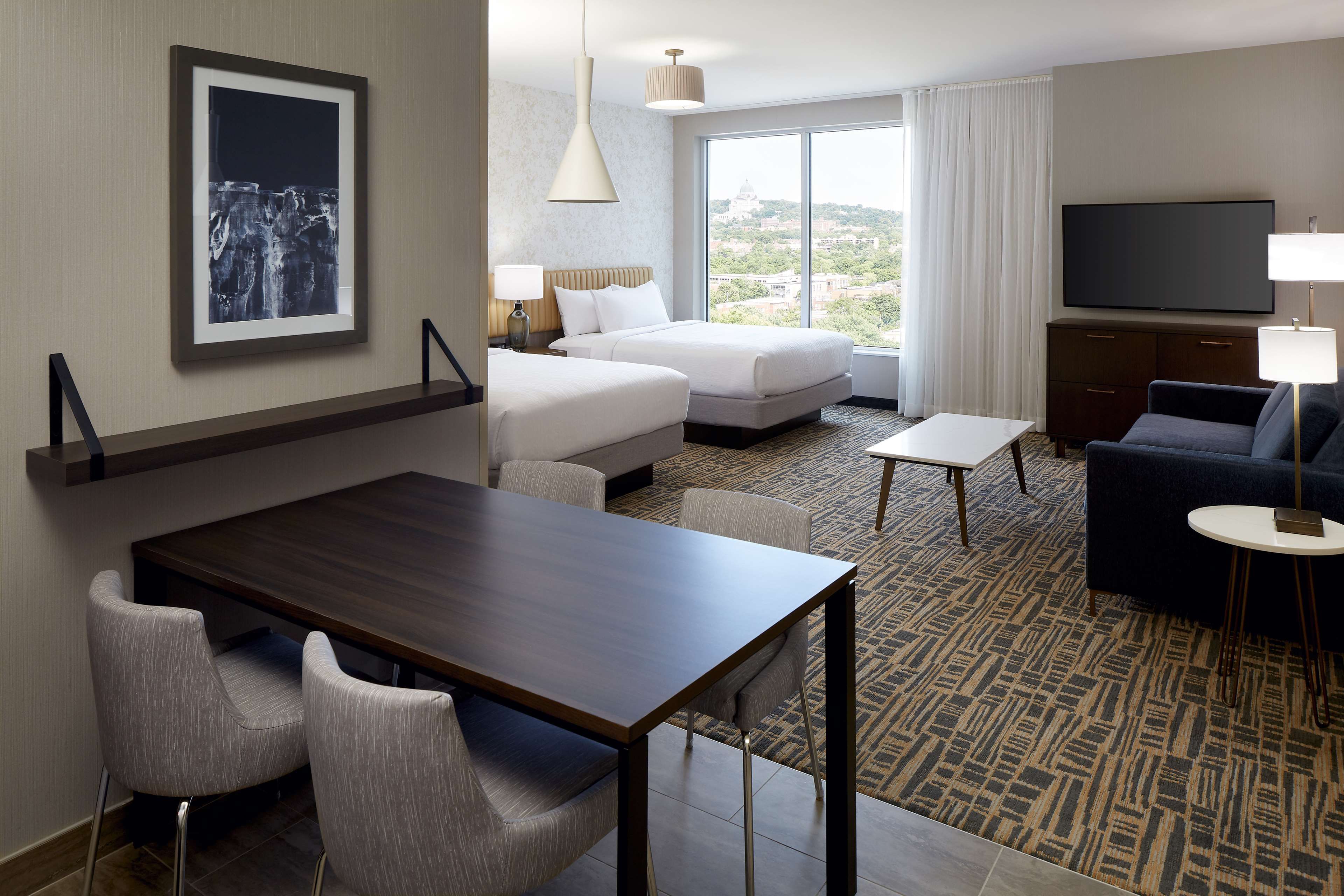 A hotel room with a bed, a TV, a dining table with chairs, and a view of the city.