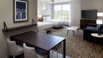 A hotel room with a bed, a TV, a dining table with chairs, and a view of the city.