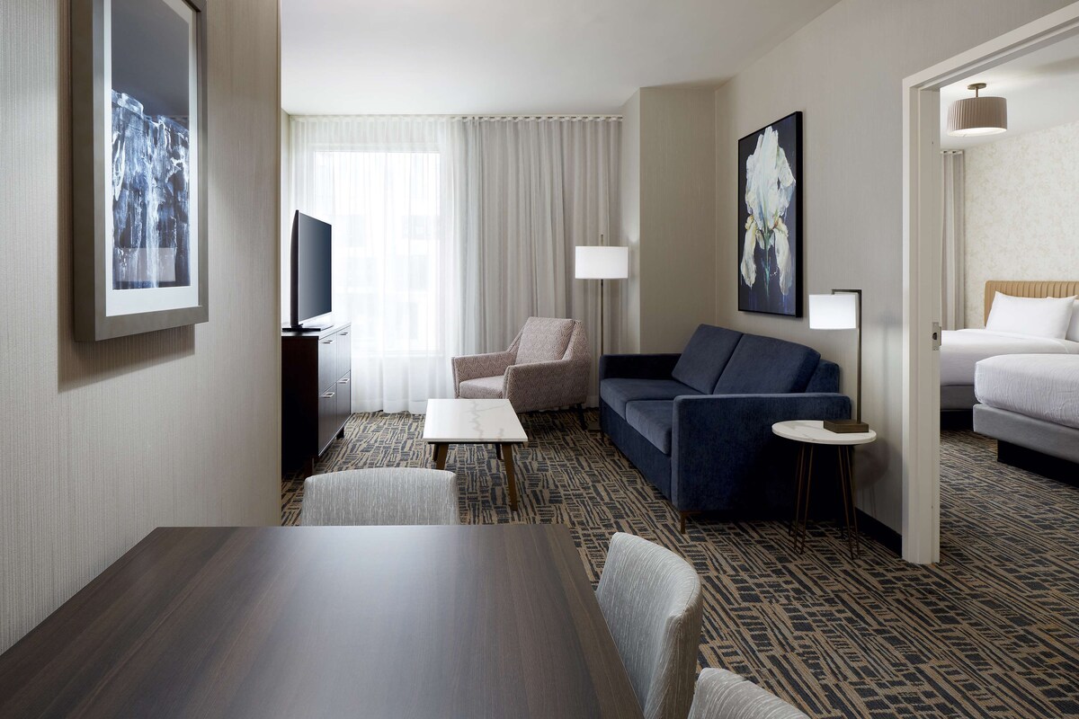 A hotel room with a dark wood table, a sofa, a chair, a TV, a lamp, and a bed.