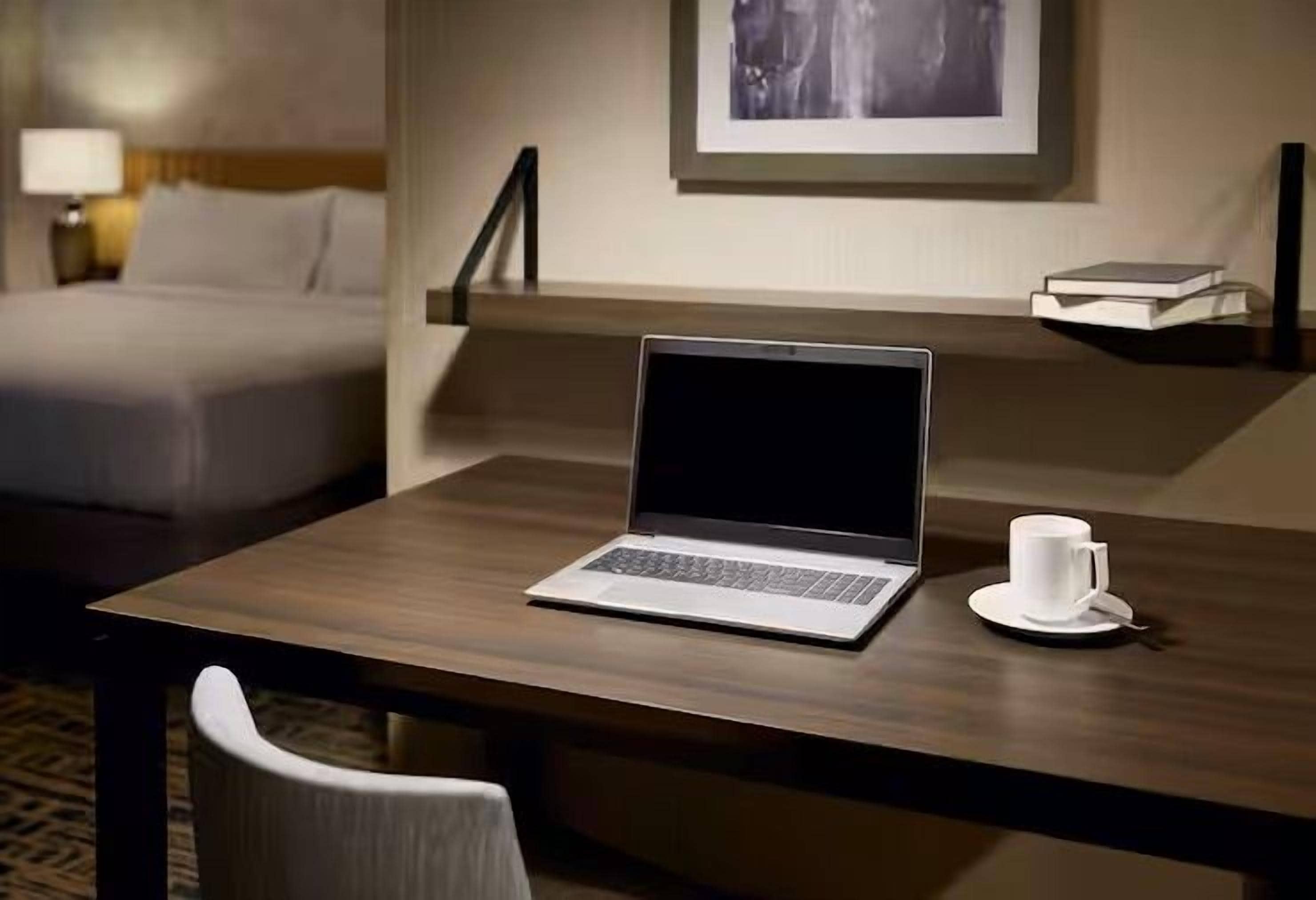 A hotel room with a bed, a desk with a laptop, a cup, and a framed picture on the wall.