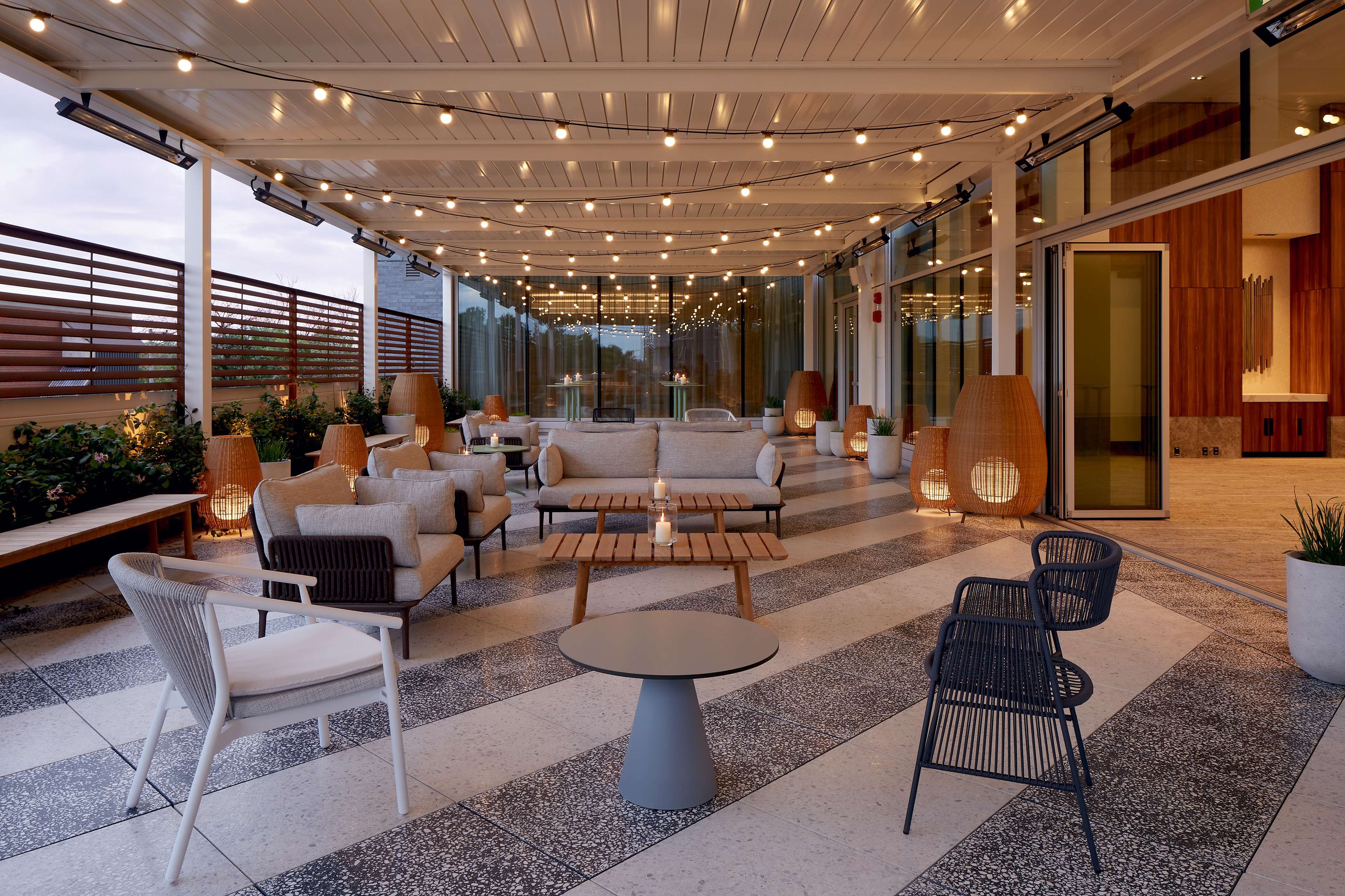 An outdoor seating area with sofas, chairs, and tables, surrounded by glass walls and string lights.