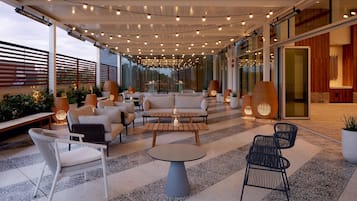 An outdoor seating area with sofas, chairs, and tables, surrounded by glass walls and string lights.