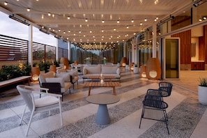 An outdoor seating area with sofas, chairs, and tables, surrounded by glass walls and string lights.