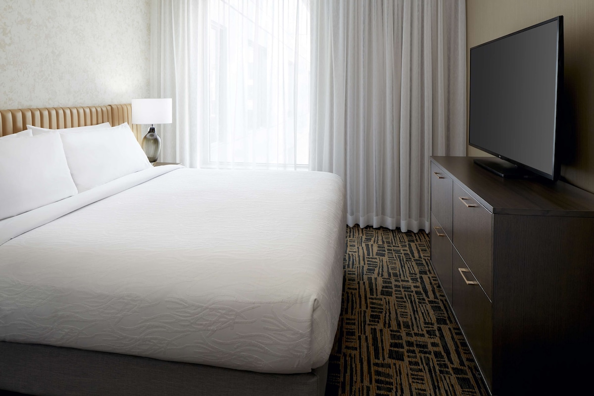 A hotel room with a large bed, a flat-screen TV, a nightstand with a lamp, and a patterned carpet.