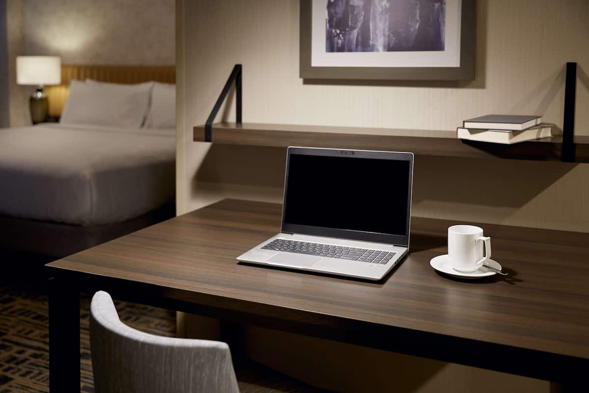 A hotel room with a wooden desk, a laptop, a white cup, and a bed in the background.