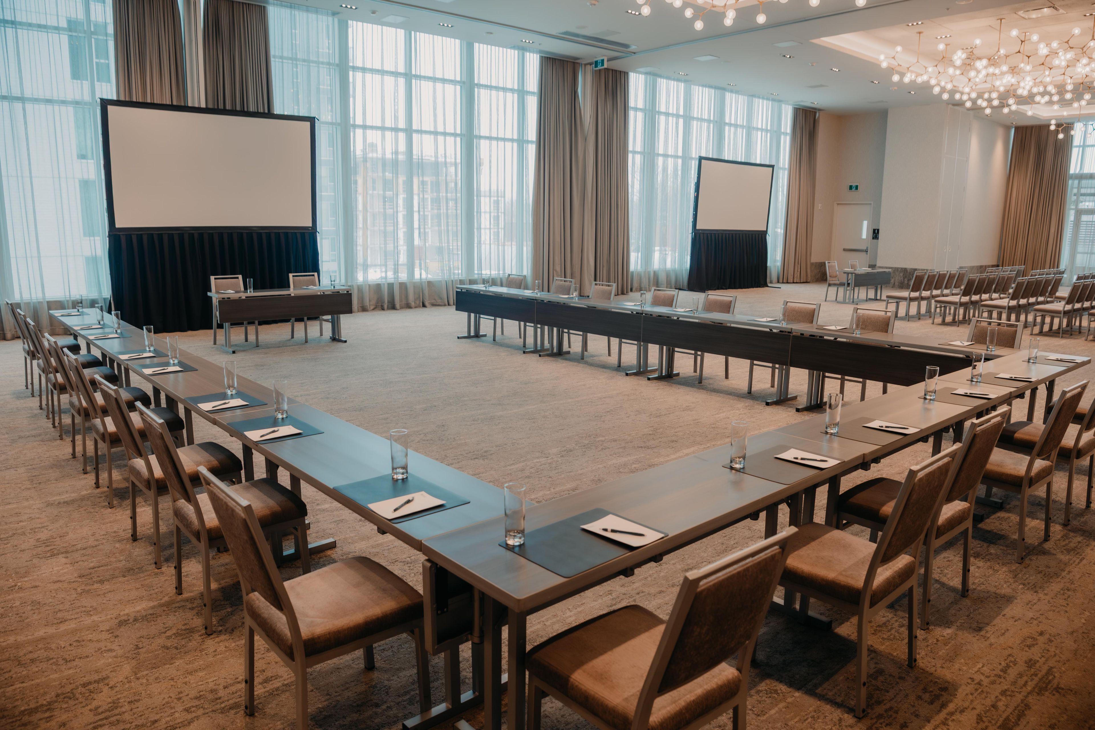 Sala de conferência com uma mesa comprida, cadeiras, tela de projetor e uma grande janela com cortinas.