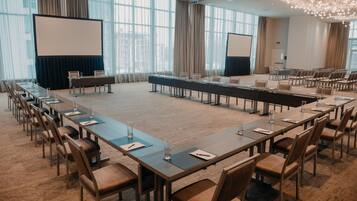 Sala de conferência com uma mesa comprida, cadeiras, tela de projetor e uma grande janela com cortinas.