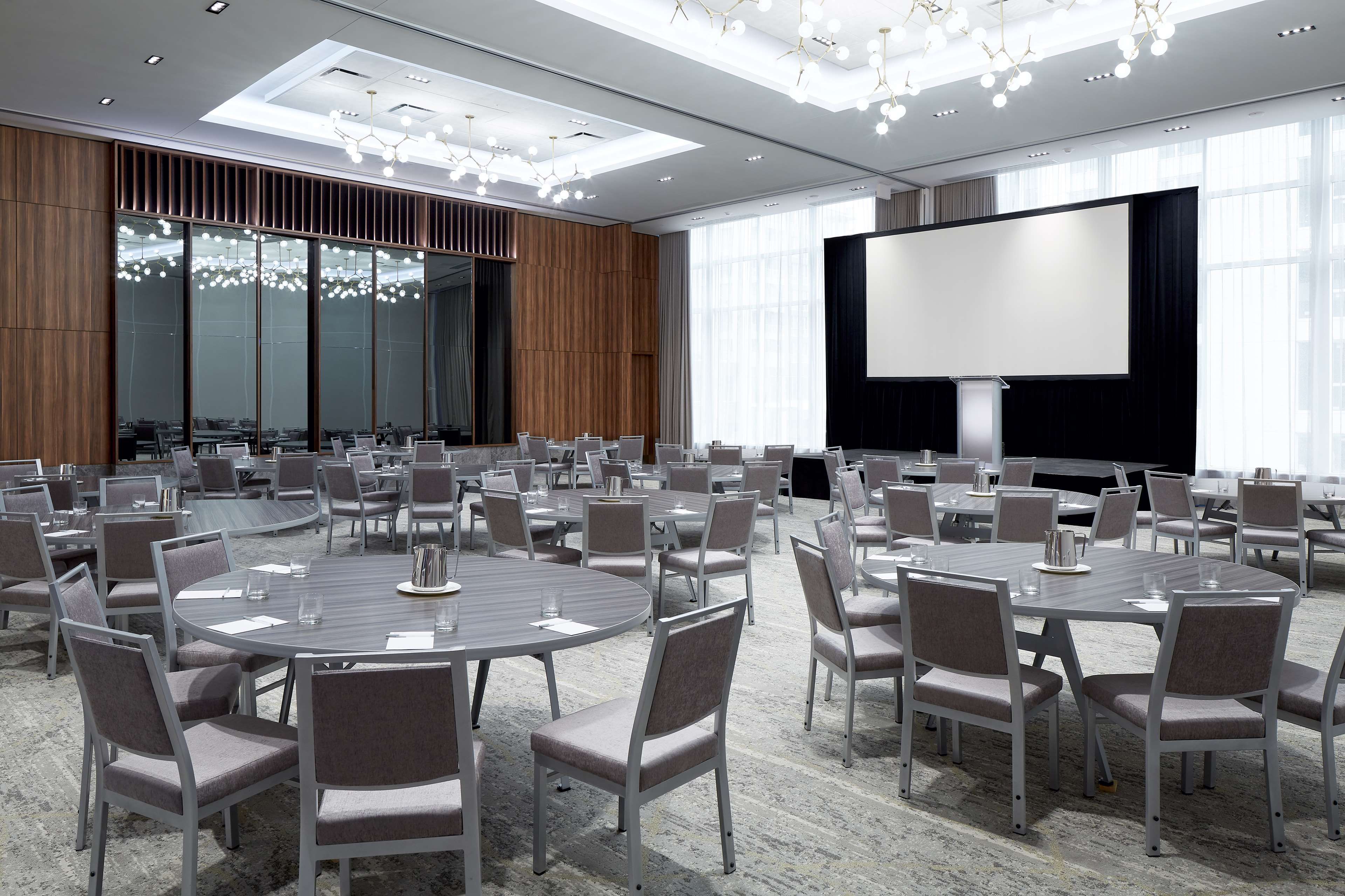Un salón de conferencias con mesas redondas, sillas, una pantalla grande y una lámpara de araña moderna.