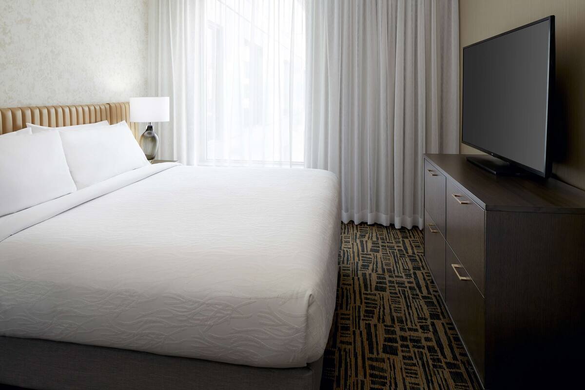 A hotel room with a large bed, a flat-screen TV, a nightstand with a lamp, and a patterned carpet.