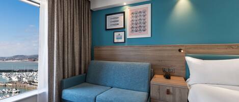 A hotel room with a sofa, a bed, a nightstand, and a view of a marina.