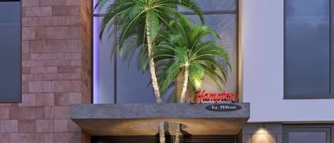 A modern building with a sign reading "Hampton by Hilton" and a large planter with two palm trees.
