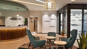 A modern reception area with a curved wooden reception desk, teal patterned chairs, and a round table. - Hampton by Hilton Torquay (Torquay)