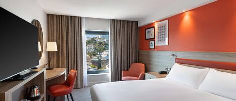 Habitación de hotel con cama, escritorio y silla, televisión y vista panorámica de la ciudad a través de la ventana.
