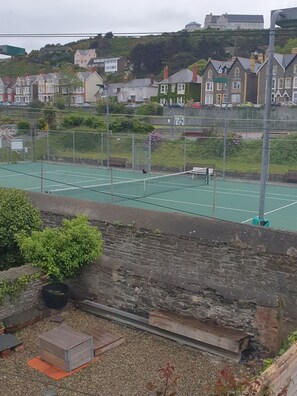 A tennis court with a net, surrounded by a fence. There are houses in the background. - Llwynygog (Aberystwyth)