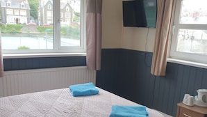Habitación de hotel con una cama grande, dos toallas azules, una mesita de noche de madera con una tetera y un televisor en la pared.