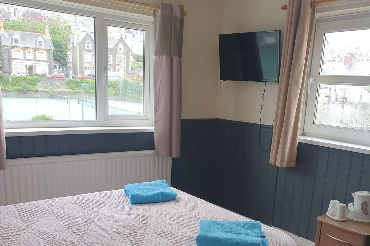 Ein Hotelzimmer mit einem großen Bett, zwei blauen Handtüchern, einem Nachttisch aus Holz mit Wasserkocher und einem an der Wand montierten Fernseher.