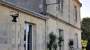 Un bâtiment en pierre de deux étages, de style architectural traditionnel, agrémenté d’une cloche, d’un lampadaire et d’un petit jardin avec des plantes et des fleurs.