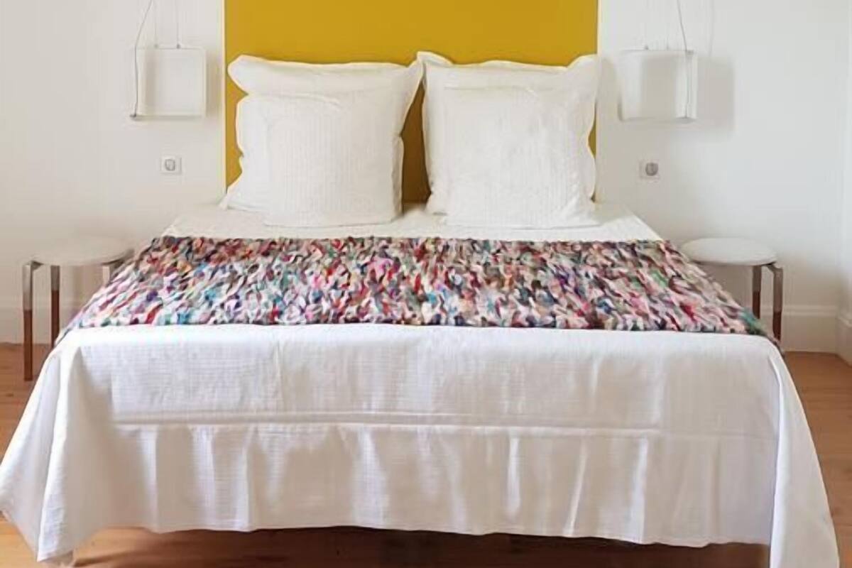 A bedroom with a bed, white bedding, a yellow accent wall, and pendant lights.