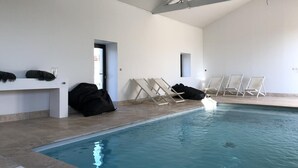 Une piscine intérieure à l’eau claire, entourée de murs blancs et dotée d’un plafond haut. Des chaises longues et un banc sont disposés le long du bord de la piscine.