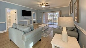 A living room with a sofa, a coffee table, a lamp, and a TV mounted on the wall. - Magnolia Place by Palmetto Vacations (Myrtle Beach)