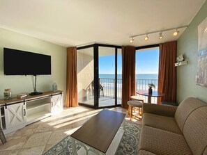 Een kamer met een flatscreen televisie, een bank, een tafel en een balkon met uitzicht op het strand.