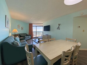 Een woonkamer met een bank, een eettafel met stoelen, een aan de muur bevestigde televisie en uitzicht op de oceaan door de schuifdeur.