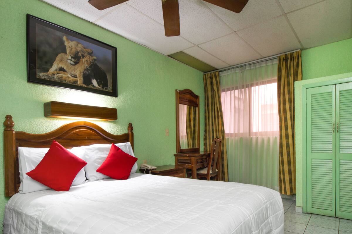 Une chambre d’hôtel avec un grand lit, une tête de lit en bois, un tableau représentant des lions, un bureau avec une chaise et une porte verte.