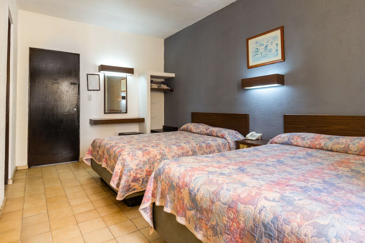 Une chambre d’hôtel avec deux lits, un ventilateur de plafond, un miroir, un tableau encadré et une petite table de chevet avec un téléphone.