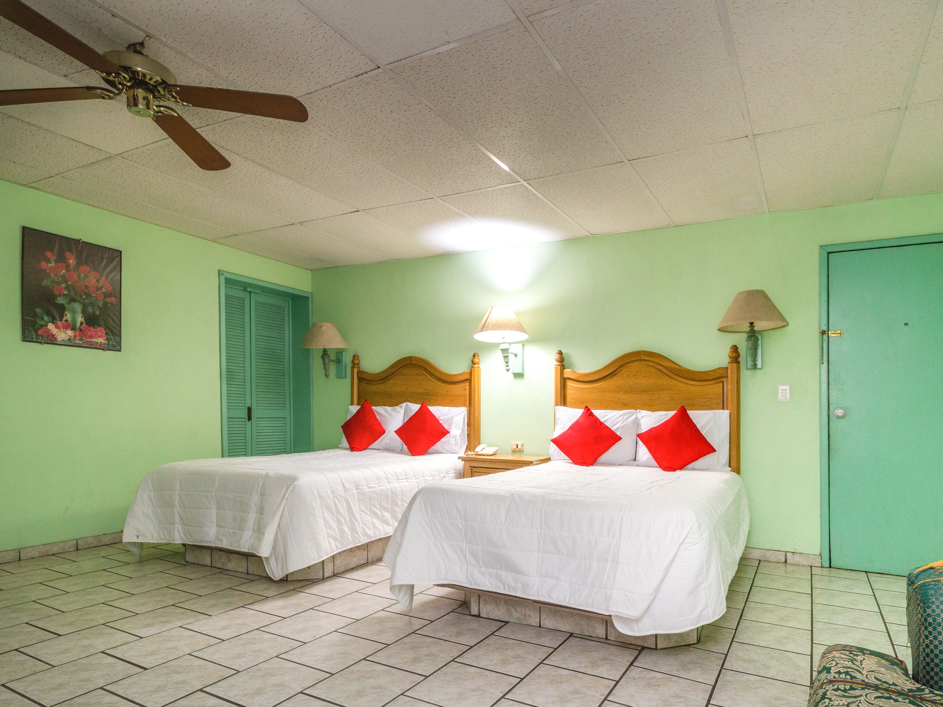Une chambre d’hôtel avec deux lits, un ventilateur de plafond, une porte verte et un tableau au mur.