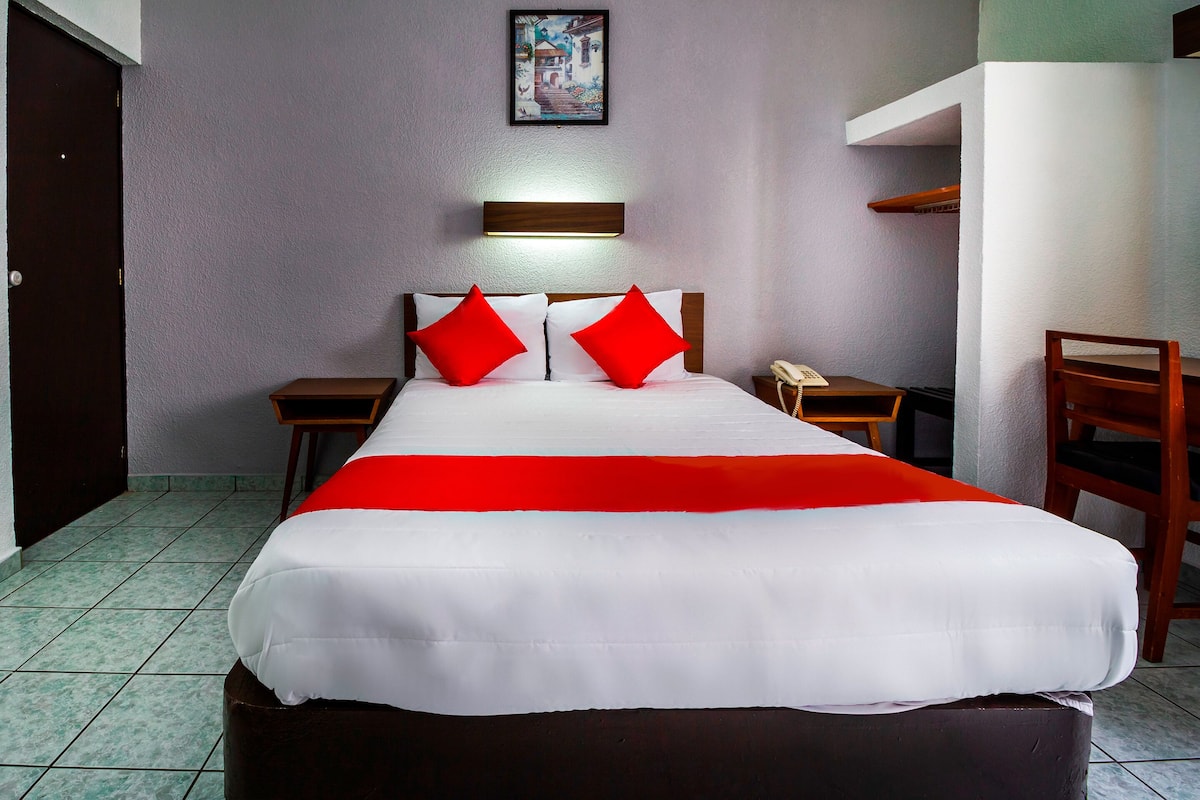Une chambre d’hôtel avec un grand lit, deux tables de chevet, une chaise en bois, une lampe fixée au mur et un tableau au-dessus du lit.