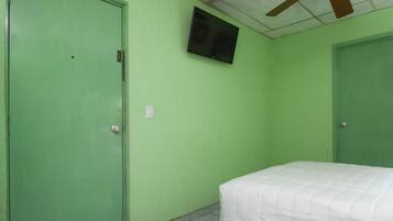 Una habitación con paredes verdes, un ventilador de techo, una cama con cubrecama blanco y un televisor montado en la pared.