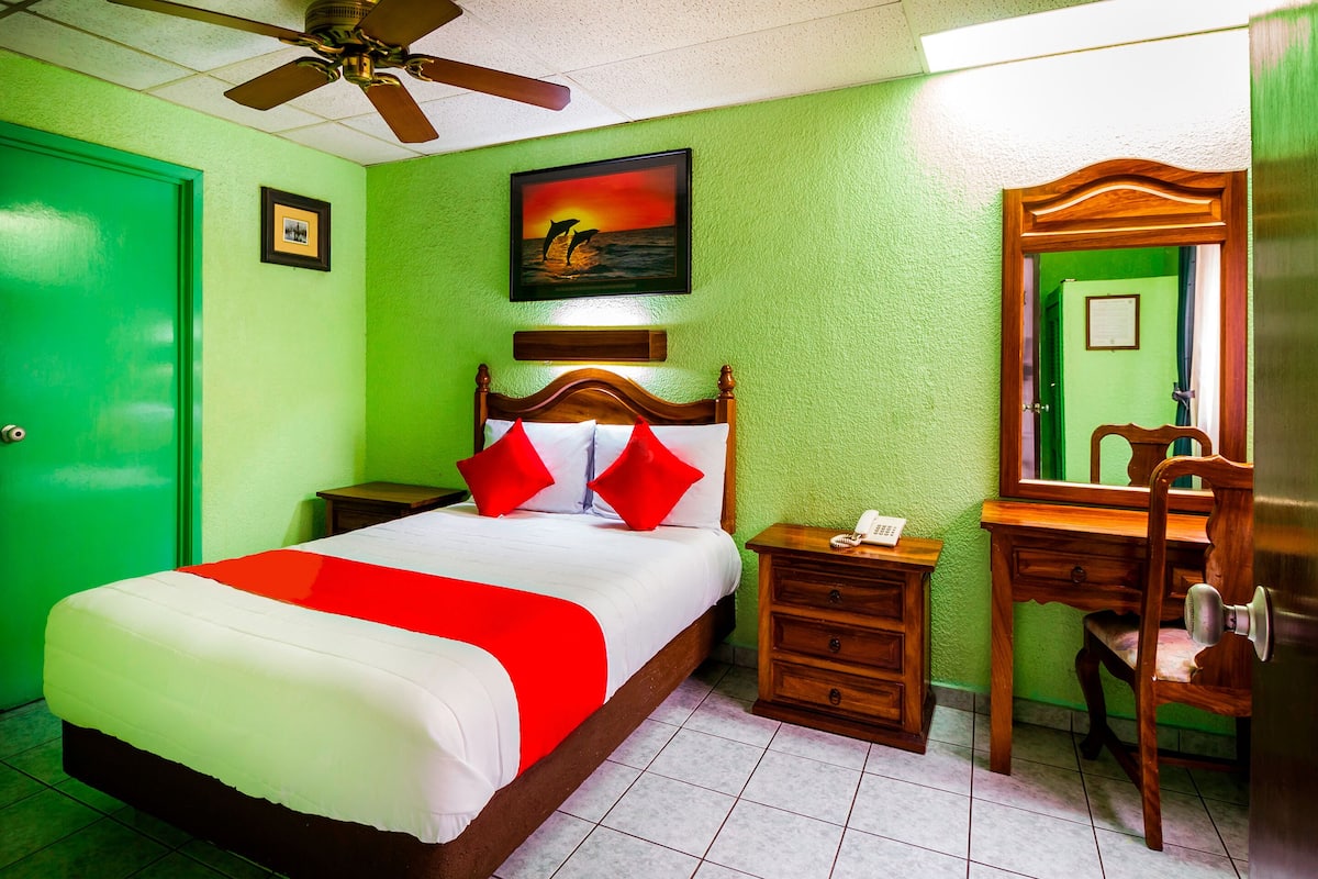 Une chambre d’hôtel avec un lit, une table de chevet en bois, un bureau avec une chaise, un ventilateur de plafond et un mur vert.