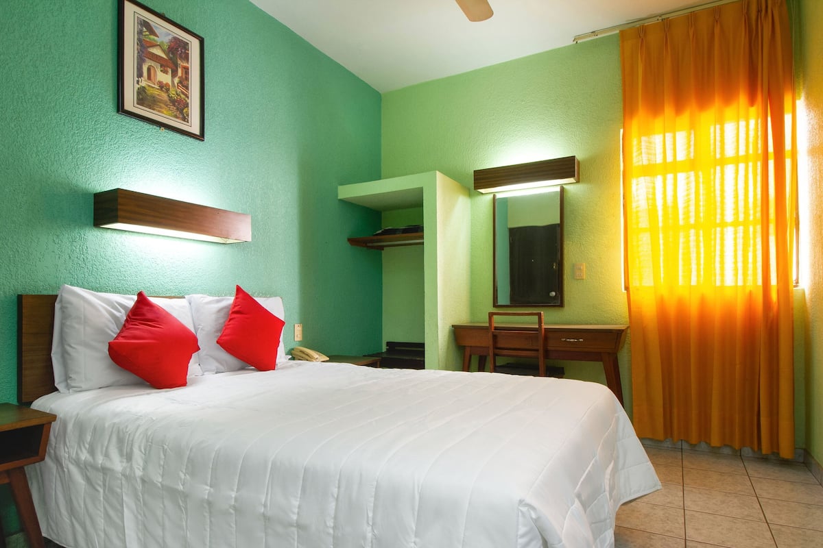 Une chambre d’hôtel équipée d’un lit, d’un bureau, d’une chaise, d’un miroir et d’un ventilateur de plafond.