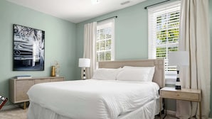 A bedroom with a large bed, two bedside tables, a nightstand, a lamp, and a window with blinds.