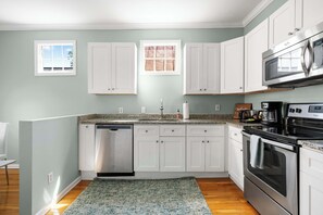 Una cocina con armarios blancos, lavavajillas, microondas y estufa. Hay dos ventanas con vistas al exterior.