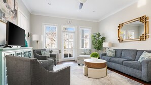 A living room with a grey sofa, a round coffee table, and a television. There are windows with blinds, a plant, and a mirror on the wall. - Rooftop Patio, Unparalleled Views in 3-story Home (Charleston)