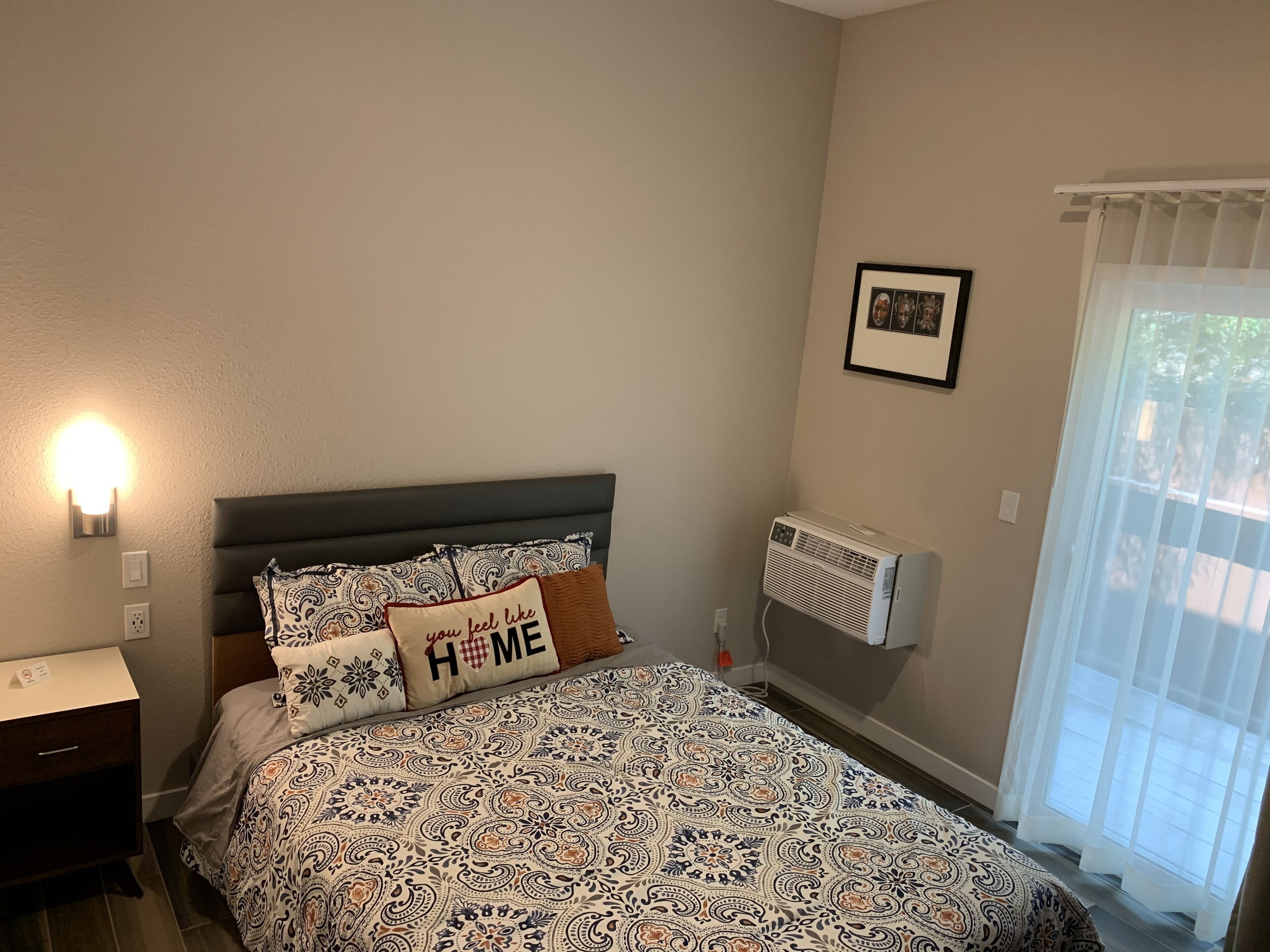 A bedroom with a patterned bedspread, a bedside table with a lamp, a wall-mounted air conditioner, and a window with sheer curtains.