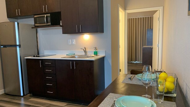 A compact kitchen with dark cabinets, a microwave, and a sink. There's a dining area with a table set for two, a glass of water, and a bowl of lemons. - Newer private apart-hotel suite in Old Town (San Diego)