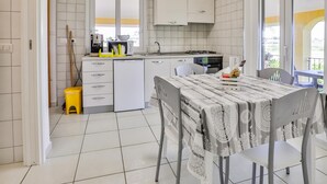 Ett modernt kök med vita skåp, ett matbord med ett mönstrat bordsduks och en utsikt genom den öppna dörren.