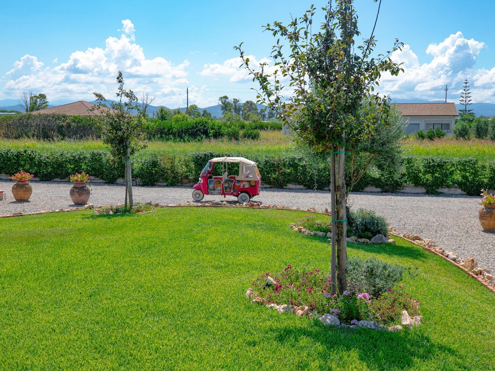 A red vehicle parked on a gravel path in a well-maintained garden with green grass, flowering plants, and a tree.